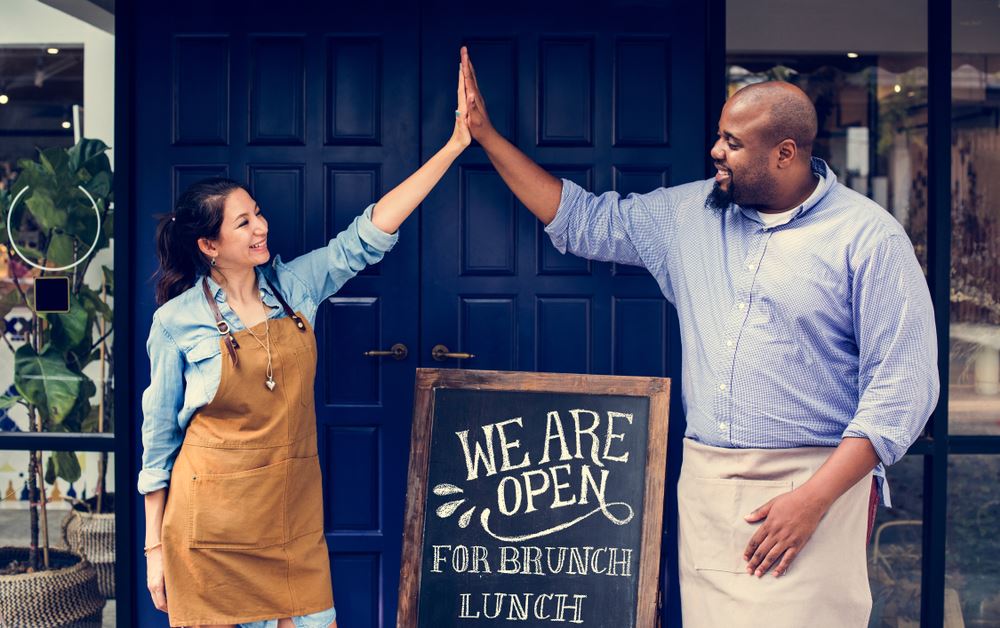Woman and man high-five in front of "We are Open" sign