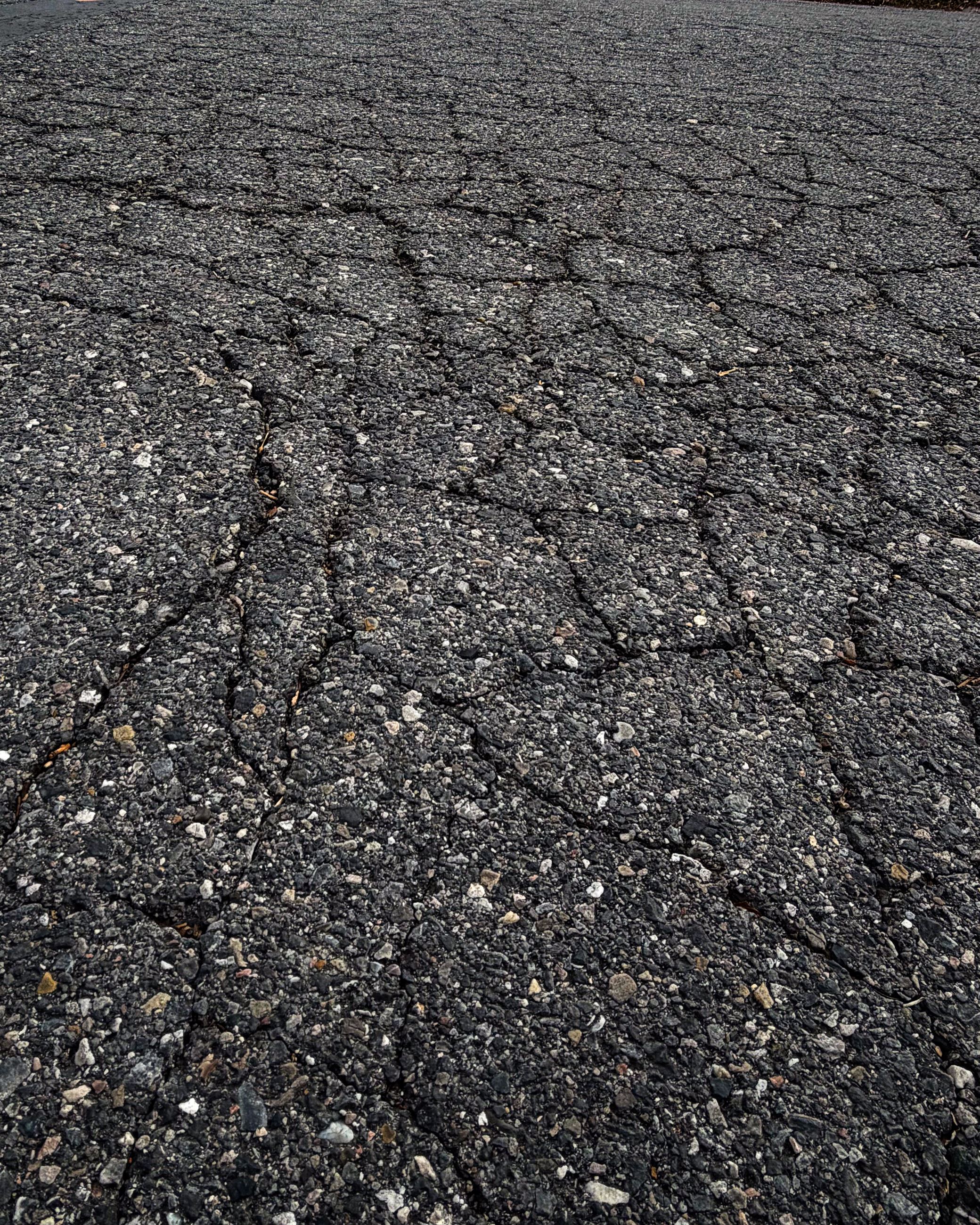 Cracked asphalt surface, showing numerous irregular fractures across a weathered, gritty pavement.