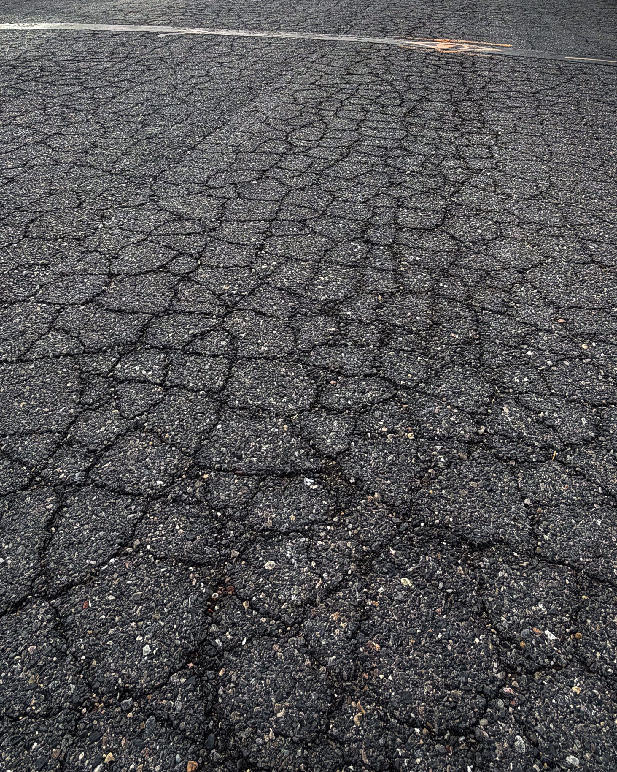 Cracked asphalt surface, showing numerous irregular fractures across a weathered, gritty pavement.